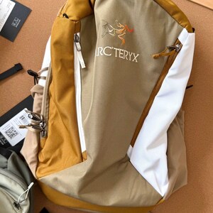 May include: Tan and white backpack with a yellow top and accents. The backpack features a zipper closure, a logo, and the text "RC'TERYX". The bag is made of a durable material and is designed for outdoor use.