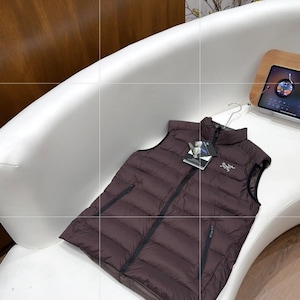 May include: A dark brown puffer vest with a black zip and a small logo on the chest. The vest is displayed on a white curved surface. The vest has a tag attached to it.