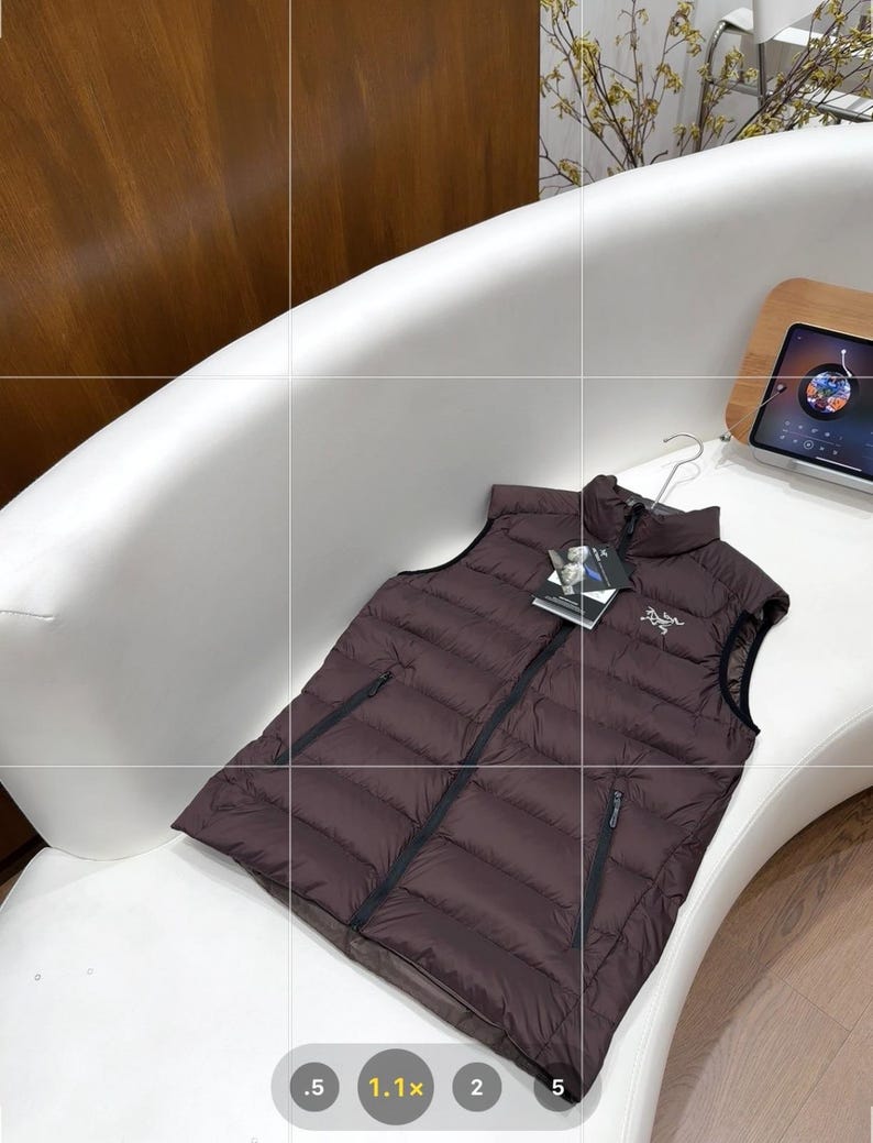 May include: A dark brown puffer vest with a black zip and a small logo on the chest. The vest is displayed on a white curved surface. The vest has a tag attached to it.