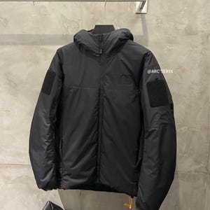 May include: Black hooded jacket with a full-length zipper, two square patches on the sleeves, and a small orange pull tab. The jacket is hanging on a metal rack against a gray wall. The text "@ARC'TERYX" is visible.