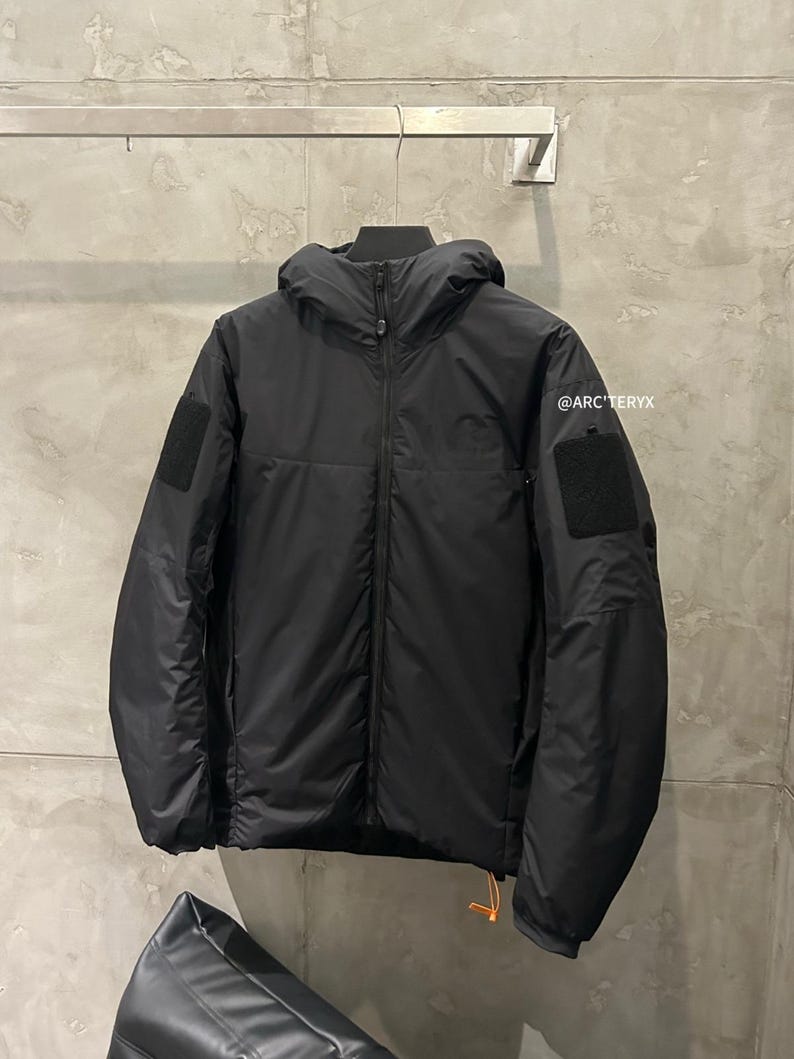 May include: Black hooded jacket with a full-length zipper, two square patches on the sleeves, and a small orange pull tab. The jacket is hanging on a metal rack against a gray wall. The text "@ARC'TERYX" is visible.