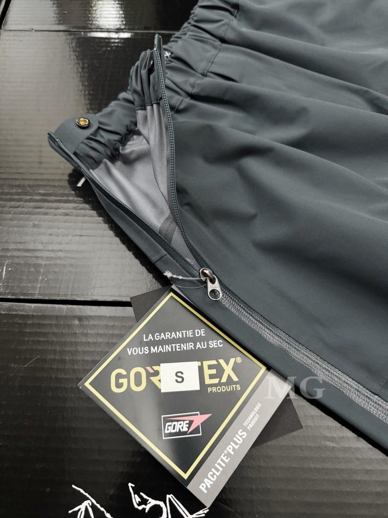 K&ouml;nnte beinhalten: Dunkelgraue wasserdichte Hose mit Rei&szlig;verschluss und elastischem Bund. Ein Etikett mit dem Text "GORE-TEX" und "PACLITE PLUS" ist angebracht. Die Hose besteht aus leichtem, strapazierf&auml;higem Material.