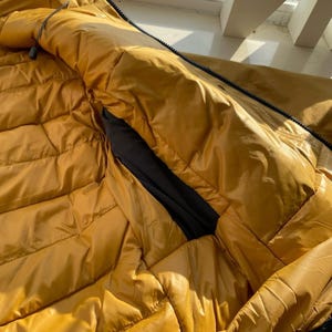 May include: Close-up of a mustard-yellow puffer jacket with a black interior pocket. The jacket has a quilted design and a black zip. The fabric has a shiny, water-resistant appearance.