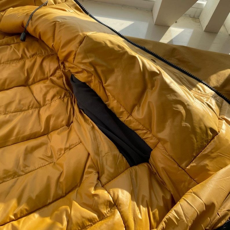 May include: Close-up of a mustard-yellow puffer jacket with a black interior pocket. The jacket has a quilted design and a black zip. The fabric has a shiny, water-resistant appearance.
