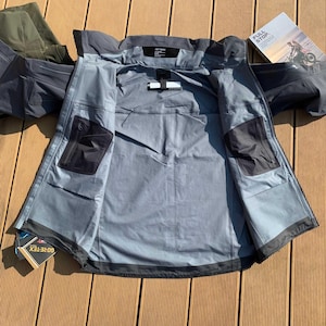 Op de afbeelding: Grijze waterdichte jas met zwarte details, open getoond om de binnenvoering en zakken te laten zien. De jas heeft een capuchon en een GORE-TEX label. Geplaatst op een houten oppervlak.