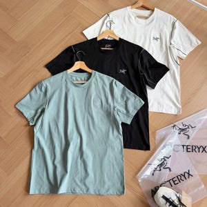 May include: Three short-sleeved t-shirts in black, white, and pale green, each with a small logo on the chest. The shirts are on wooden hangers. Two clear plastic bags with a logo and a pair of white and grey trainers are also visible.