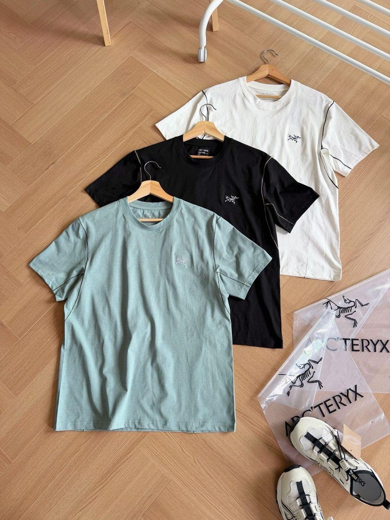 May include: Three short-sleeved t-shirts in black, white, and pale green, each with a small logo on the chest. The shirts are on wooden hangers. Two clear plastic bags with a logo and a pair of white and grey trainers are also visible.