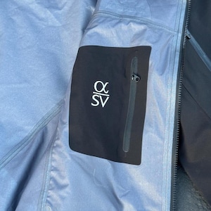 May include: Close-up of the interior of a black and gray jacket. A black pocket with the white text "&alpha; SV" is visible. The jacket has a zipper and a light blue lining.