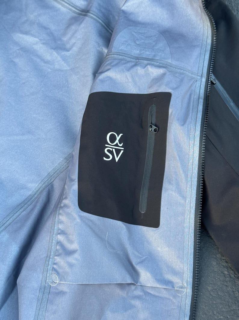 May include: Close-up of the interior of a black and gray jacket. A black pocket with the white text "&alpha; SV" is visible. The jacket has a zipper and a light blue lining.