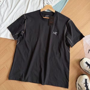 May include: Black short-sleeved t-shirt with a small logo on the chest and a tag. The shirt has light-coloured stitching along the seams. Additional white and light blue t-shirts are visible in the background.