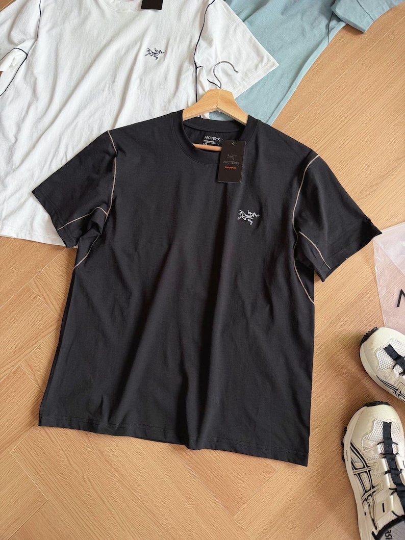 May include: Black short-sleeved t-shirt with a small logo on the chest and a tag. The shirt has light-coloured stitching along the seams. Additional white and light blue t-shirts are visible in the background.