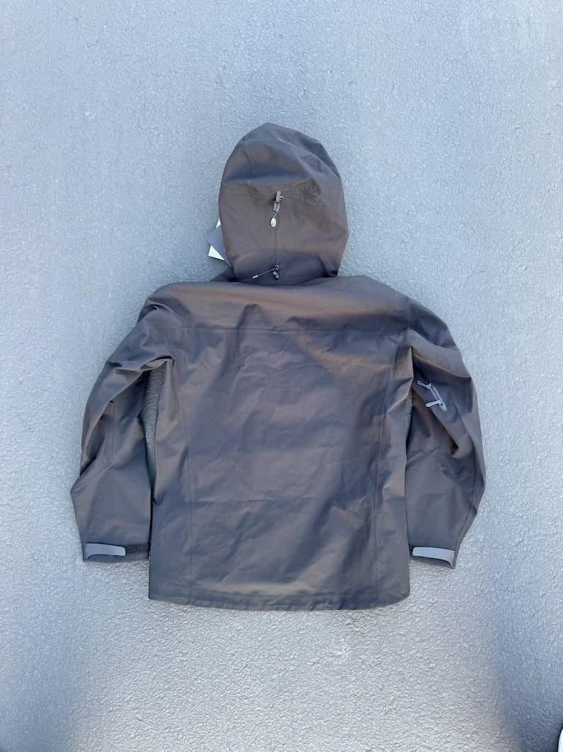 May include: A dark gray hooded jacket with a zipper closure and a small logo on the sleeve. The jacket has a hood with a drawstring and adjustable cuffs. The jacket is made of a lightweight, waterproof material.
