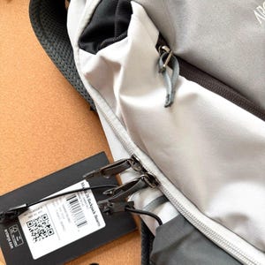 May include: Close-up of a gray and white backpack with black accents. The backpack has a zippered compartment and a black tag attached. The tag has text and a QR code. The backpack is made of a durable material and has a padded shoulder strap.