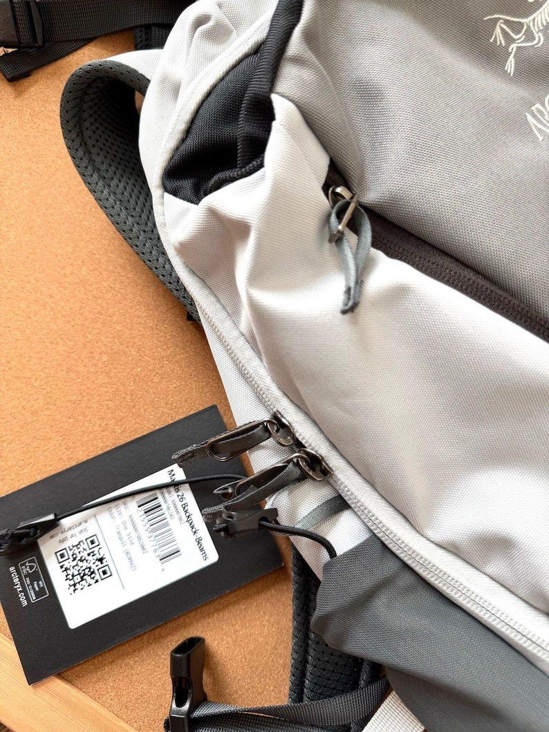 May include: Close-up of a gray and white backpack with black accents. The backpack has a zippered compartment and a black tag attached. The tag has text and a QR code. The backpack is made of a durable material and has a padded shoulder strap.