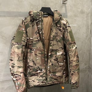 May include: A camouflage-patterned jacket with a hood, hanging on a metal rack. The jacket features a full-length zipper, multiple pockets, and the brand name "Arc'teryx" on the chest. The color palette includes shades of green, brown, and tan.