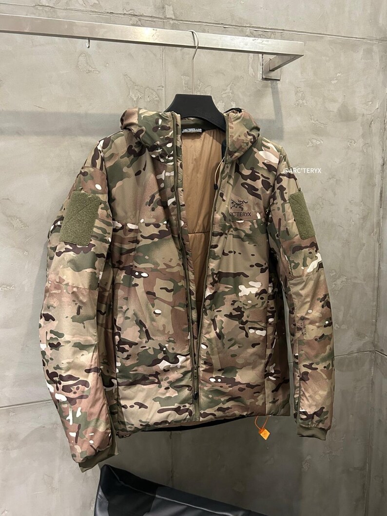 May include: A camouflage-patterned jacket with a hood, hanging on a metal rack. The jacket features a full-length zipper, multiple pockets, and the brand name "Arc'teryx" on the chest. The color palette includes shades of green, brown, and tan.