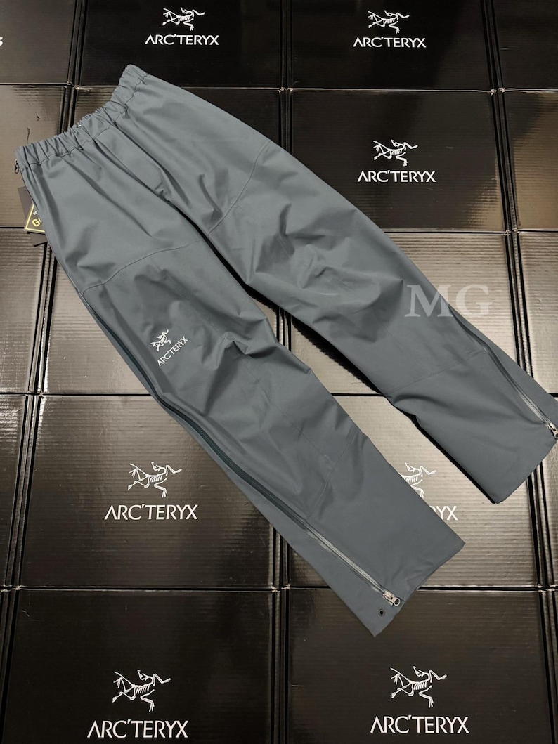 K&ouml;nnte beinhalten: Graue Arc'teryx-Regenhose mit elastischem Bund und Rei&szlig;verschlusstaschen. Die Hose hat das Arc'teryx-Logo auf dem linken Bein und Rei&szlig;verschl&uuml;sse an den Kn&ouml;cheln. Hergestellt aus leichtem, wasserdichtem Material.