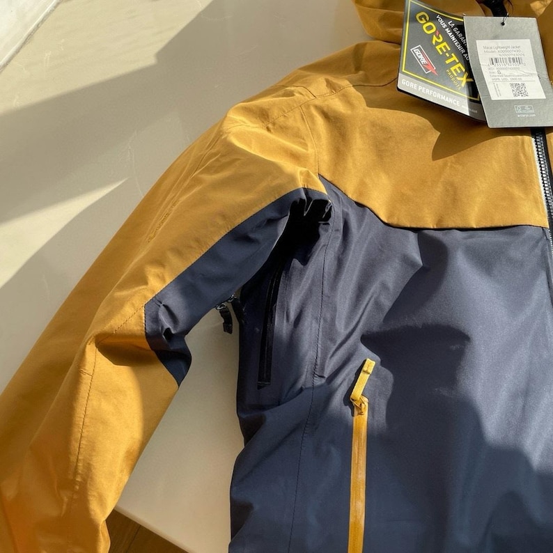 May include: A mustard yellow and navy blue outdoor jacket with a hood. The jacket has a zip closure and multiple pockets. The GORE-TEX label is visible. The jacket is designed for outdoor activities.