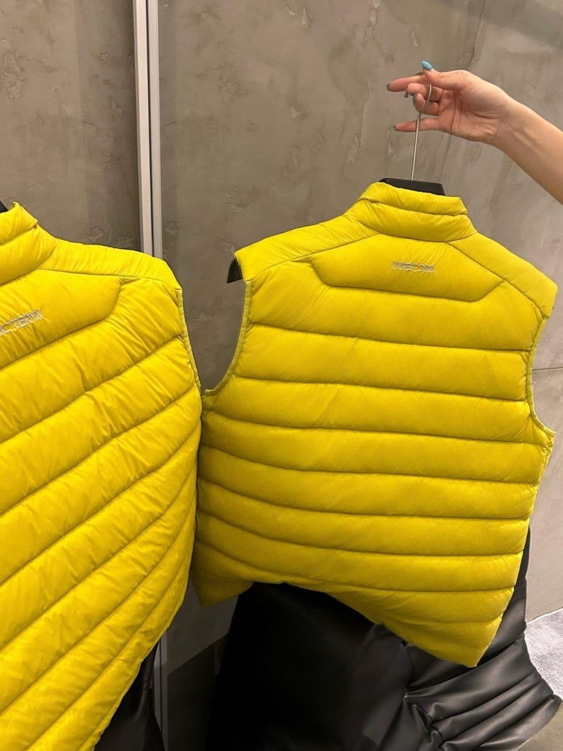 May include: Two bright yellow puffer vests are displayed. The vests have a high collar and a horizontal quilted pattern. The vests are hanging on black hangers. The brand name is visible on the back of one of the vests.