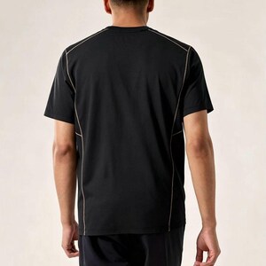 May include: Black t-shirt with tan stitching detail. The short-sleeved shirt is visible from the back. The model is wearing black shorts. The shirt has a crew neck. The seams are highlighted with a contrasting colour.