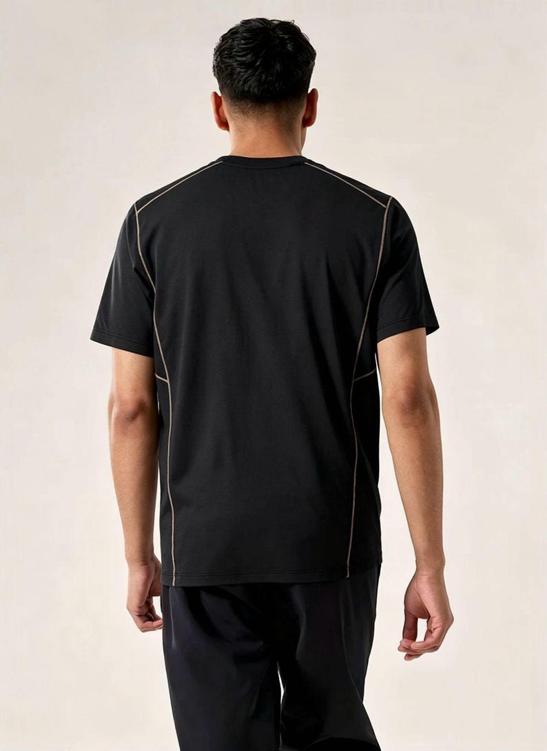 May include: Black t-shirt with tan stitching detail. The short-sleeved shirt is visible from the back. The model is wearing black shorts. The shirt has a crew neck. The seams are highlighted with a contrasting colour.