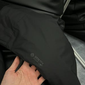 May include: Black jacket with the text "GORE-TEX INFINIUM" printed on the sleeve. The jacket appears to be made of a smooth, matte material. The image shows a close-up of the jacket's sleeve.