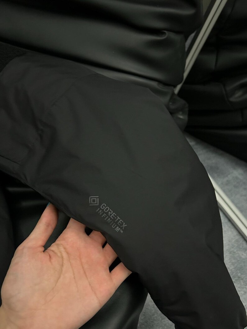 May include: Black jacket with the text "GORE-TEX INFINIUM" printed on the sleeve. The jacket appears to be made of a smooth, matte material. The image shows a close-up of the jacket's sleeve.