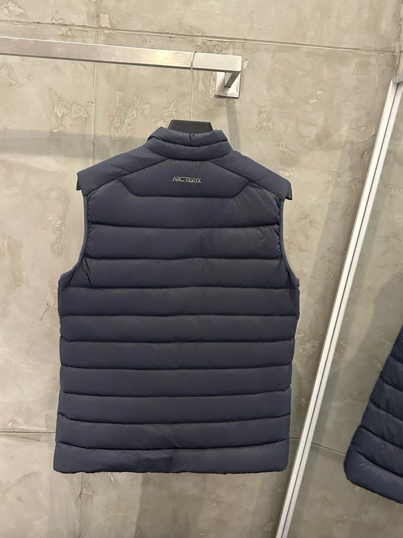 May include: A navy blue puffer vest with horizontal quilted stitching. The vest has a stand-up collar and the brand name "ARCTERYX" is printed on the upper back. The vest is hanging on a black hanger.
