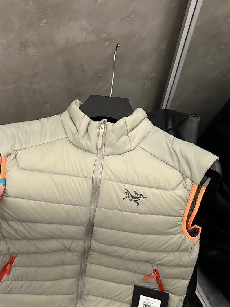 May include: A light grey puffer vest with a full-length zipper and a small logo on the chest. The vest has orange trim around the armholes and a small orange zipper pull. The vest is hanging on a black hanger.