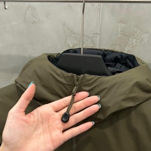 May include: An olive green jacket with a high collar and a black interior. The jacket has a zipper closure with a black pull tab. The jacket is hanging on a metal hanger.
