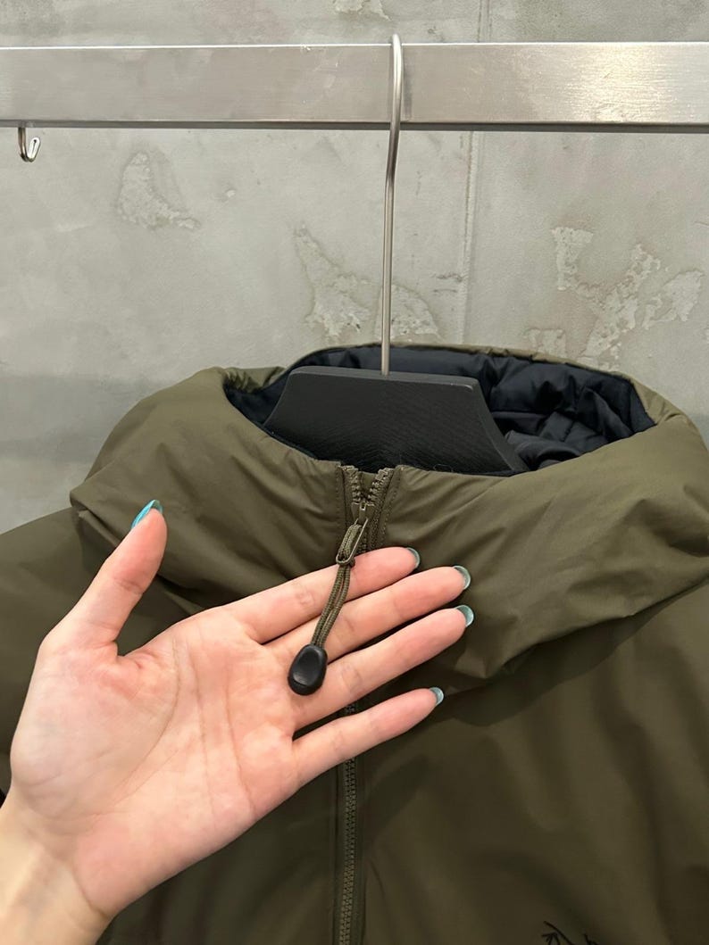 May include: An olive green jacket with a high collar and a black interior. The jacket has a zipper closure with a black pull tab. The jacket is hanging on a metal hanger.