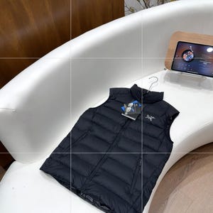 May include: A black puffer vest with a full-length zip and a high collar. The vest is displayed on a white curved surface. A tag with the brand logo is attached. The vest is designed for warmth and outdoor activities.