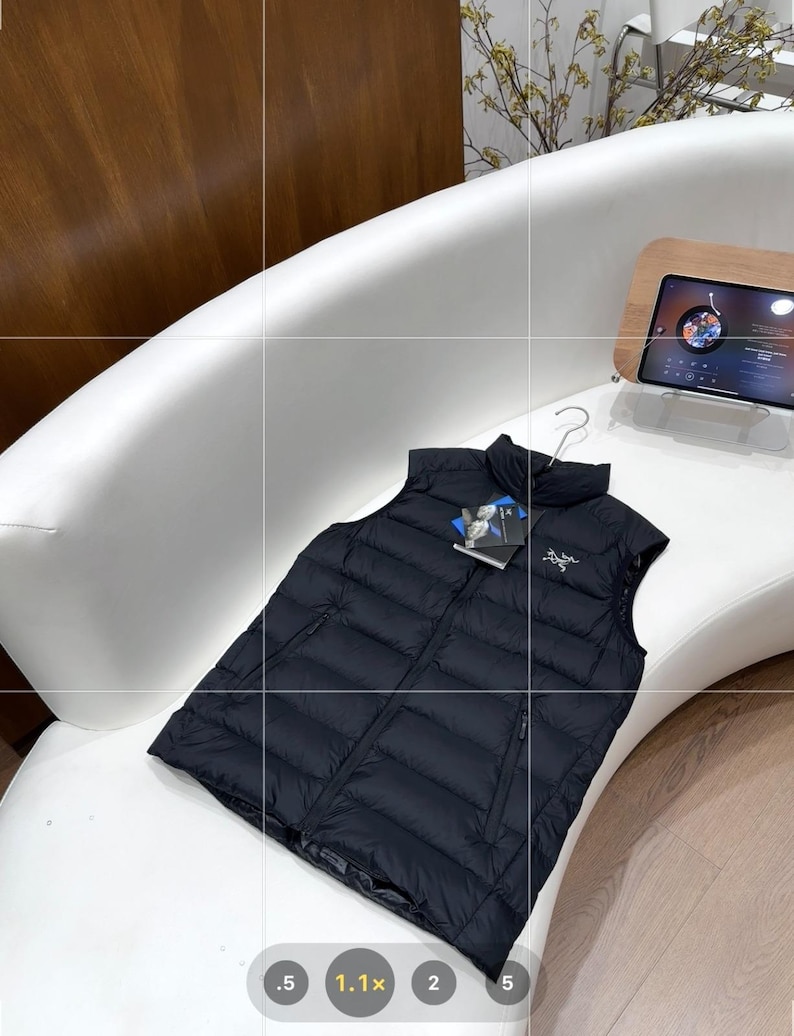 May include: A black puffer vest with a full-length zip and a high collar. The vest is displayed on a white curved surface. A tag with the brand logo is attached. The vest is designed for warmth and outdoor activities.