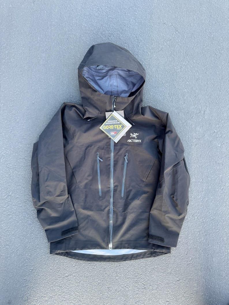 May include: A dark gray hooded jacket with a full-length zipper and multiple pockets. The jacket features the "GORE-TEX" and "Arc'teryx" logos. The jacket is designed for outdoor activities and protection from the elements.