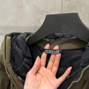 May include: An olive green Arc'teryx jacket with a black interior lining. The jacket is hanging on a black hanger. The tag reads "Arc'teryx" and "L/G".