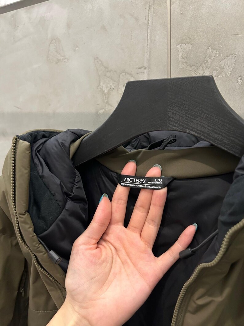 May include: An olive green Arc'teryx jacket with a black interior lining. The jacket is hanging on a black hanger. The tag reads "Arc'teryx" and "L/G".