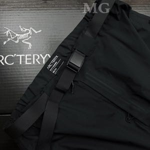 May include: Black athletic pants with a black adjustable belt and buckle. The pants have a small zipper pocket and the brand name "Arc'teryx" is visible. The pants are made of a lightweight material.