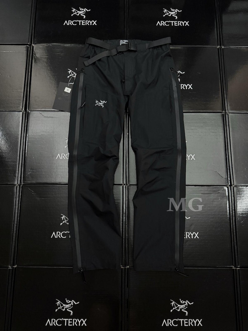 May include: Black outdoor pants with a belt, featuring the Arc'teryx logo. The pants have a streamlined design with vertical stripes down the sides and zippers at the ankles. The pants are displayed against a backdrop of stacked black boxes.