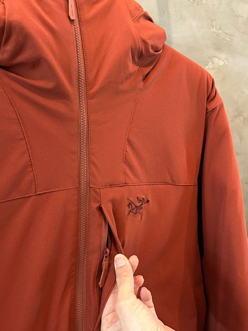 Op de afbeelding: Roestkleurige jas met capuchon, een ritssluiting over de hele lengte en een klein logo op de borst. De jas heeft een zak met ritssluiting en een gestroomlijnd ontwerp, geschikt voor buitenactiviteiten.