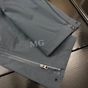 K&ouml;nnte beinhalten: Nahaufnahme einer dunkelgrauen Jacke mit Rei&szlig;verschluss und den Buchstaben "MG" auf dem Stoff. Die Jacke hat ein schlankes, modernes Design mit Fokus auf Funktionalit&auml;t und Stil. Der Rei&szlig;verschluss ist silberfarben.