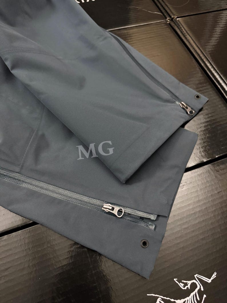 K&ouml;nnte beinhalten: Nahaufnahme einer dunkelgrauen Jacke mit Rei&szlig;verschluss und den Buchstaben "MG" auf dem Stoff. Die Jacke hat ein schlankes, modernes Design mit Fokus auf Funktionalit&auml;t und Stil. Der Rei&szlig;verschluss ist silberfarben.