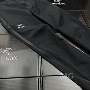 K&ouml;nnte beinhalten: Schwarze wasserdichte Hose mit wei&szlig;em Arc'teryx-Logo. Die Hose hat einen Kordelzugbund, einen Beinabschluss mit Rei&szlig;verschluss und einen reflektierenden Streifen. Auf schwarzen Boxen mit Arc'teryx-Logo platziert.