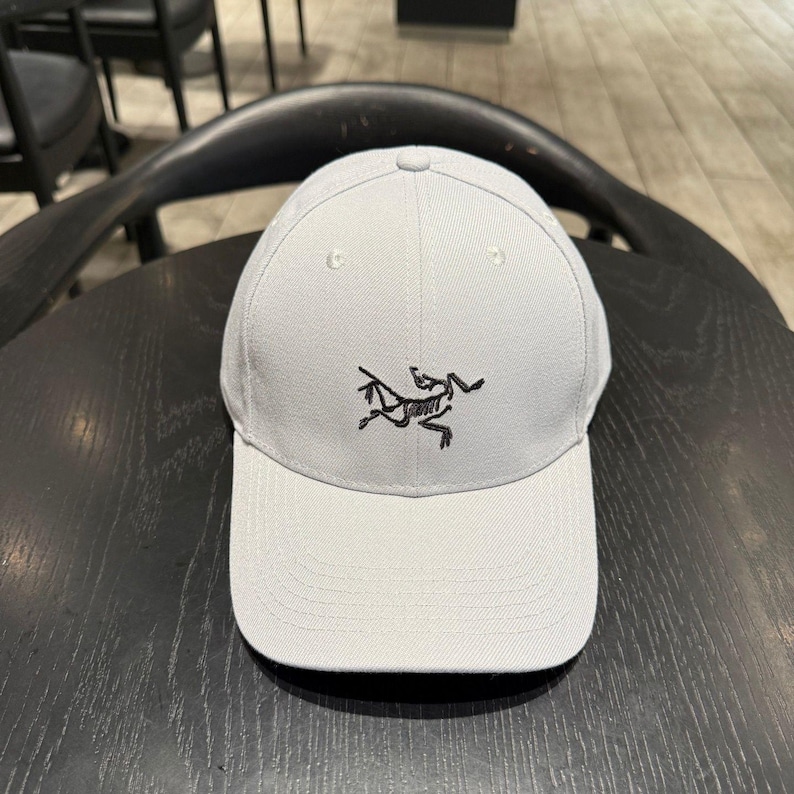 Puede incluir: Gorra de b&eacute;isbol gris claro con visera curva. La gorra presenta un logotipo negro bordado en la parte delantera. La gorra est&aacute; hecha de una tela lisa y tiene una corona estructurada.