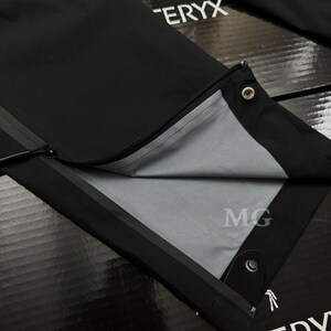 May include: Close-up of black outdoor pants with a zipper detail. The pants have a gray interior lining and a snap closure at the bottom. The brand name is visible in white text.