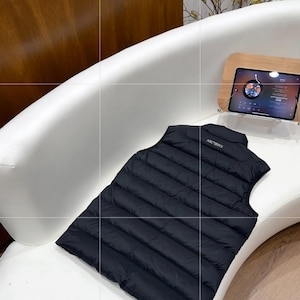 May include: A black, quilted vest with the brand name "Arc'teryx" on the back, placed on a white curved surface. The vest is a solid, dark colour and appears to be made of a lightweight material. A tablet is visible in the background.