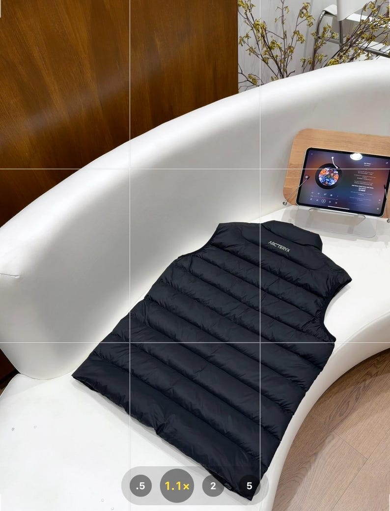 May include: A black, quilted vest with the brand name "Arc'teryx" on the back, placed on a white curved surface. The vest is a solid, dark colour and appears to be made of a lightweight material. A tablet is visible in the background.