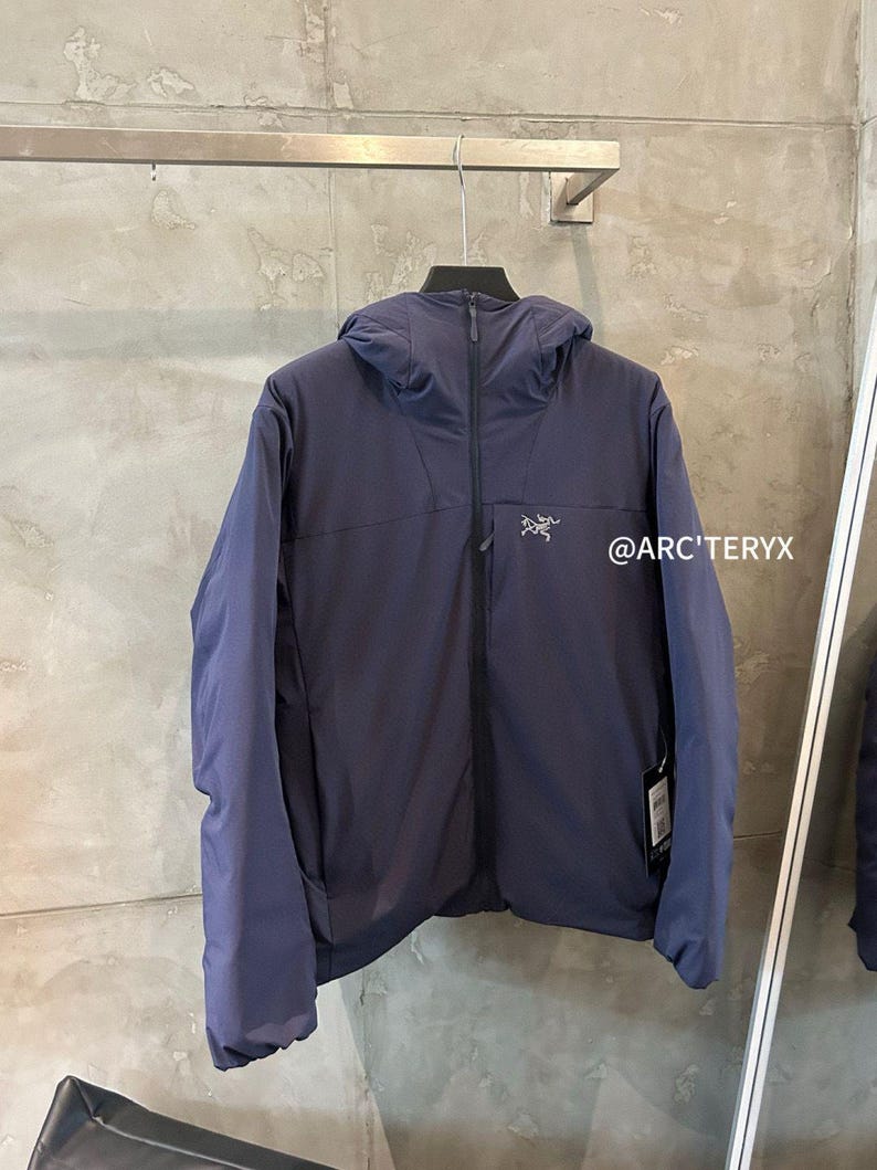 Op de afbeelding: Marineblauwe jas met capuchon, ritssluiting aan de voorkant en het merklogo op de borst. De jas hangt aan een metalen rek tegen een grijze muur. De tekst "@ARC'TERYX" is zichtbaar.