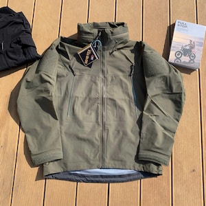 Op de afbeelding: Olijfgroene jas met een hoge kraag en ritssluiting. De jas heeft een GORE-TEX-label en patches op de schouders. Links is een zwart kledingstuk zichtbaar en rechts een boek met de titel "FULL STOP".