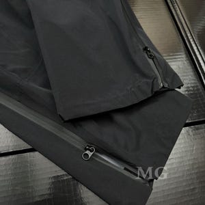 K&ouml;nnte beinhalten: Schwarze wasserdichte Hose mit Rei&szlig;verschl&uuml;ssen an den Seiten. Die Hose besteht aus einem glatten, matten Material. Das Bild zeigt eine Nahaufnahme der Hose, die die Rei&szlig;verschl&uuml;sse und die Textur des Stoffes hervorhebt. Die Hose ist gefaltet und zeigt die Innen- und Au&szlig;enseite.