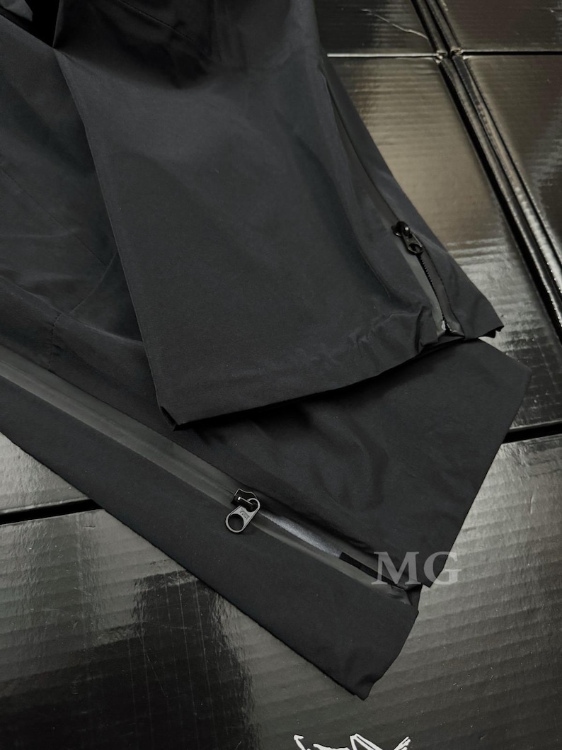 K&ouml;nnte beinhalten: Schwarze wasserdichte Hose mit Rei&szlig;verschl&uuml;ssen an den Seiten. Die Hose besteht aus einem glatten, matten Material. Das Bild zeigt eine Nahaufnahme der Hose, die die Rei&szlig;verschl&uuml;sse und die Textur des Stoffes hervorhebt. Die Hose ist gefaltet und zeigt die Innen- und Au&szlig;enseite.